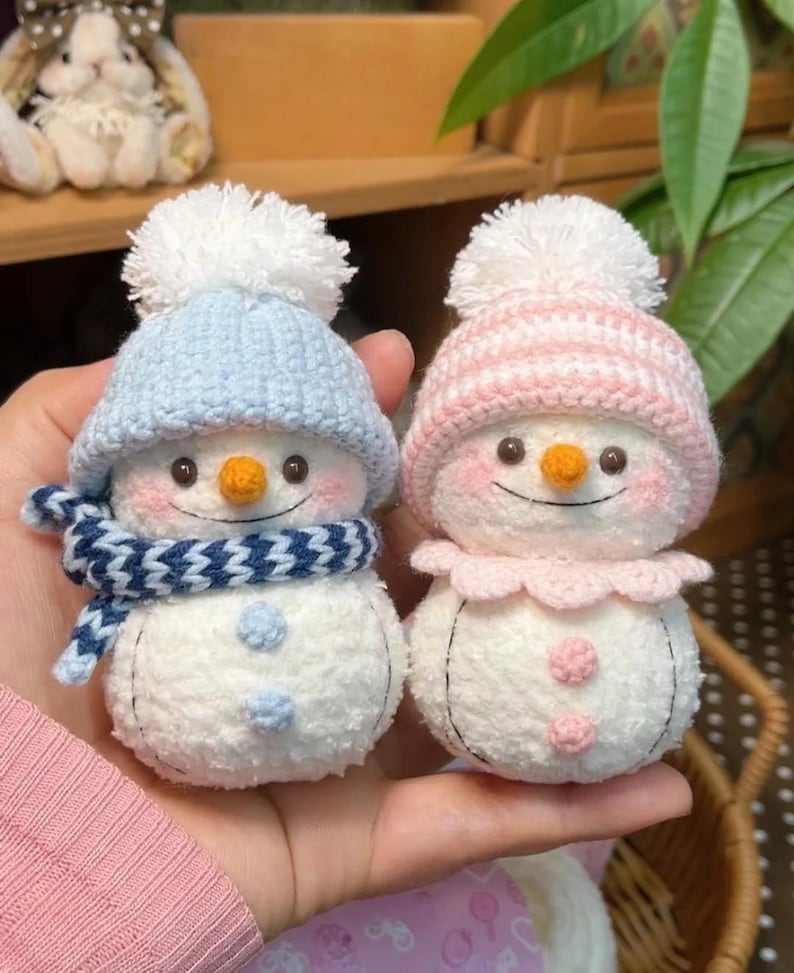May include: Two handmade snowman ornaments. One wears a blue knit hat and scarf, the other a pink hat and collar. Both have orange carrot noses, rosy cheeks, and stitched smiles. They are made of white plush material.