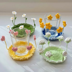 May include: Four circular crochet coasters with attached vertical stems featuring small fabric flowers and hearts. One coaster is green with white daisies, one is yellow with pink hearts, one is blue with yellow stars, and one is light green with white roses.