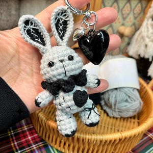 Może przedstawiać: Ręcznie robiony brelok do kluczy z królikiem z szydełka w kolorze szarym i czarnym. Królik ma czarną muszkę, detale w kształcie serca i czarny charms w kształcie serca. Brelok jest przymocowany do srebrnego pierścienia.