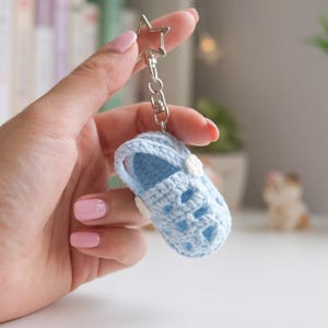Op de afbeelding: Een lichtblauwe gehaakte babyschoen-sleutelhanger met een stervormige metalen sluiting. Het miniatuurschoentje is gemaakt met een getextureerd garenpatroon en bevat kleine witte ronde accenten aan de zijkanten, bevestigd aan een zilverkleurige ketting.