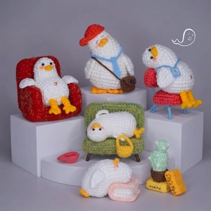 May include: A collection of five white crocheted duck figures with orange beaks and feet. One sits in a red armchair, one wears a red cap and brown bag, one sits on a red chair, one lies on a green sofa with a yellow bag, and one rests on a book near a cactus labeled MATH.