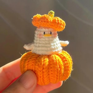 May include: A handmade crochet pumpkin-shaped figure with a white body, orange pumpkin hat, and a small green stem. The figure has a cute face with pink cheeks and small black eyes. The figure is a whimsical, handcrafted item.