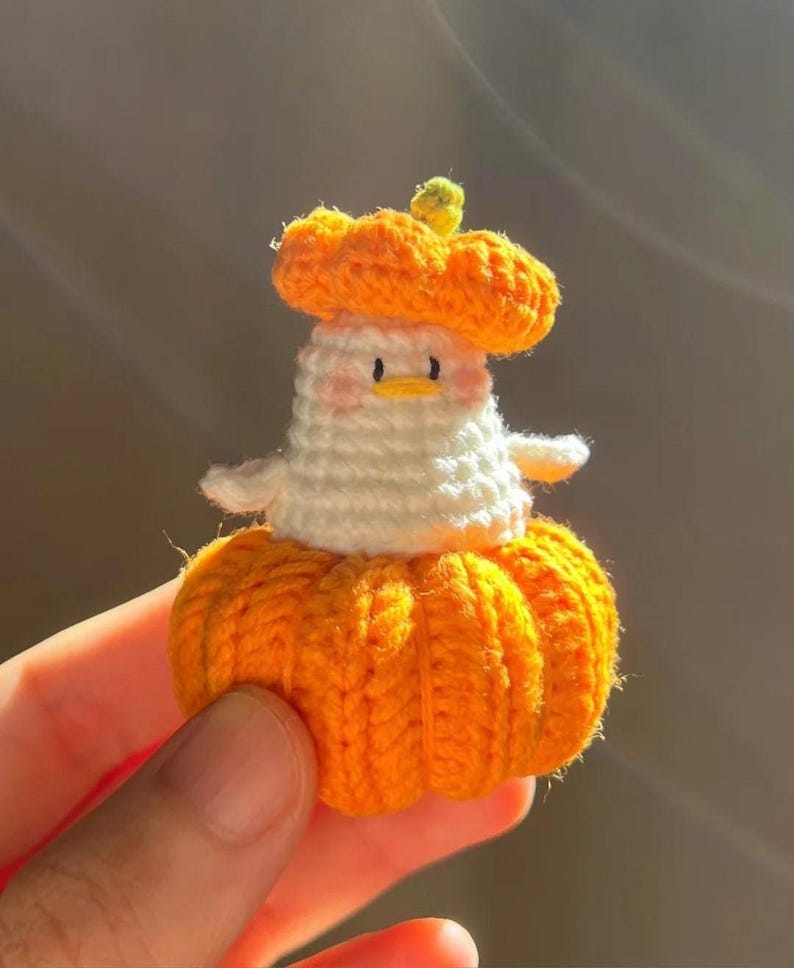 May include: A handmade crochet pumpkin-shaped figure with a white body, orange pumpkin hat, and a small green stem. The figure has a cute face with pink cheeks and small black eyes. The figure is a whimsical, handcrafted item.