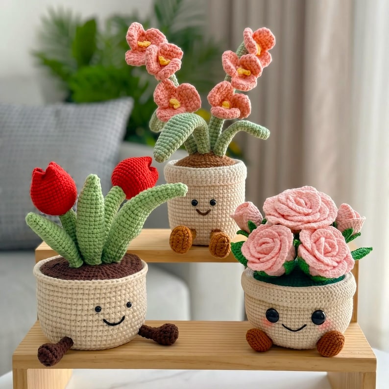 May include: Three crocheted potted plants with smiling faces. One pot holds two red tulips with green leaves, another holds a tall pink flower stalk, and the third contains a cluster of light pink roses. Each beige pot has brown feet.