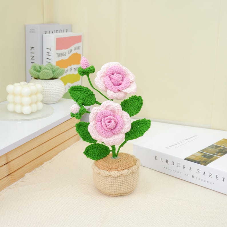 May include: A handcrafted crochet flower arrangement featuring two pink roses with green leaves and buds, planted in a beige pot. The flowers have a light pink center and a darker pink outer layer. The pot has a decorative lace trim.