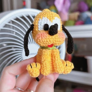 Puede incluir: Una figura de perro Pluto hecha a mano con ganchillo. El perro amarillo tiene orejas negras, una nariz negra y una lengua roja. El perro tiene detalles blancos en la cara y un collar verde. La figura es un artículo lindo y artesanal.