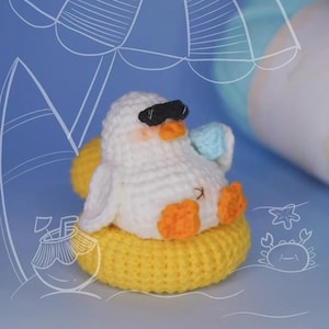 May include: A white crocheted duckling with orange feet and beak, wearing black sunglasses and holding a small blue drink. The duckling is sitting inside a bright yellow crocheted inner tube float, set against a blue background with white line art.