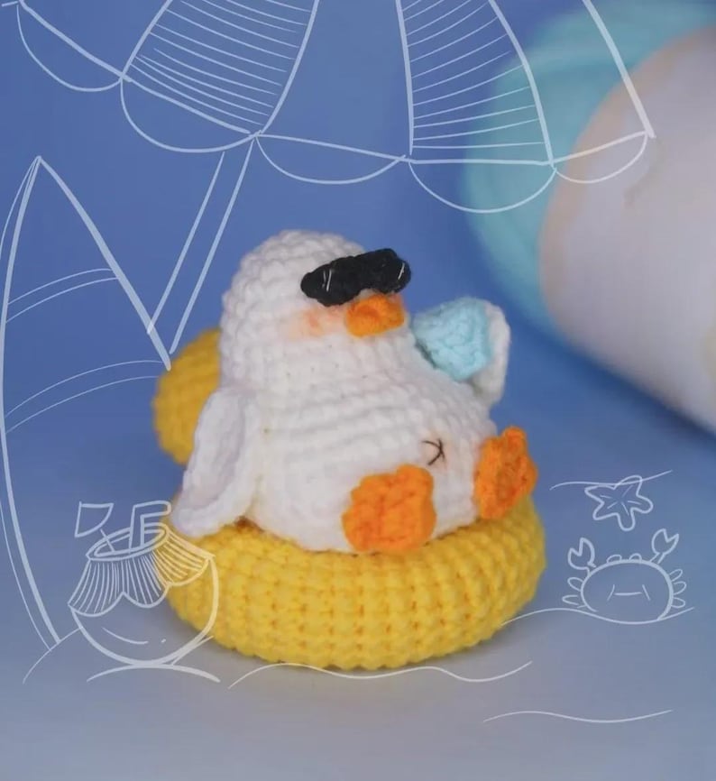 May include: A white crocheted duckling with orange feet and beak, wearing black sunglasses and holding a small blue drink. The duckling is sitting inside a bright yellow crocheted inner tube float, set against a blue background with white line art.
