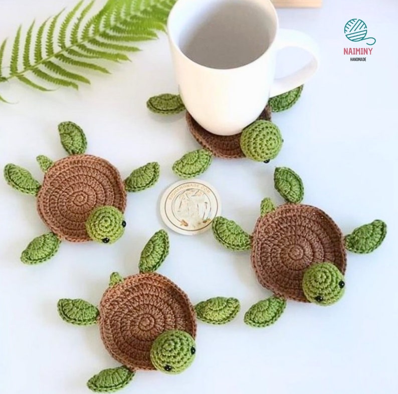 May include: Four crocheted turtle coasters in brown and green. The coasters are shaped like turtles with brown shells and green heads, legs, and tails. The coasters are sitting on a white surface.