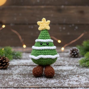 May include: A crocheted Christmas tree with a yellow star on top. The tree is green with white stripes and has small black eyes and brown feet. The tree is standing on a wooden surface with snow.