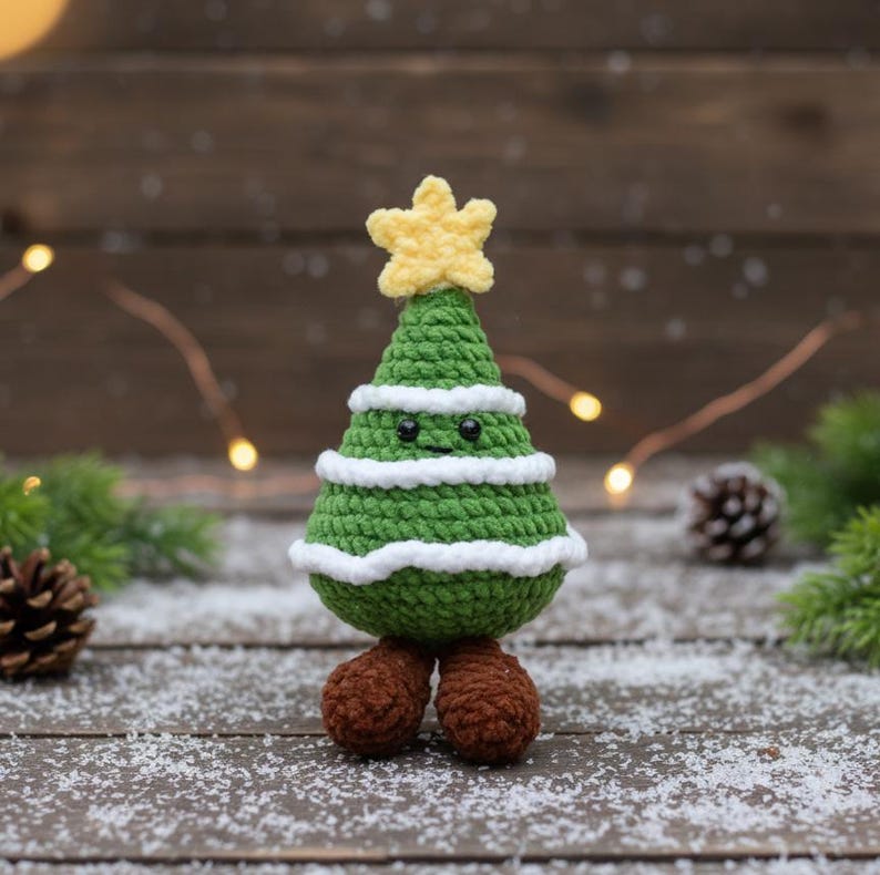 May include: A crocheted Christmas tree with a yellow star on top. The tree is green with white stripes and has small black eyes and brown feet. The tree is standing on a wooden surface with snow.