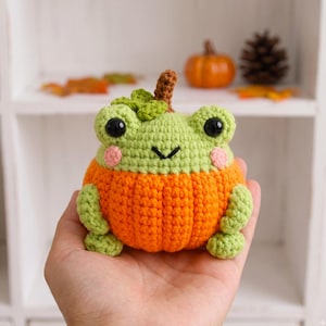 May include: A crocheted frog plushie with a light green head and a bright orange pumpkin-shaped body. The frog features black safety eyes, a small stitched mouth, pink cheeks, and a small brown stem on top of its head with a green leaf accent.