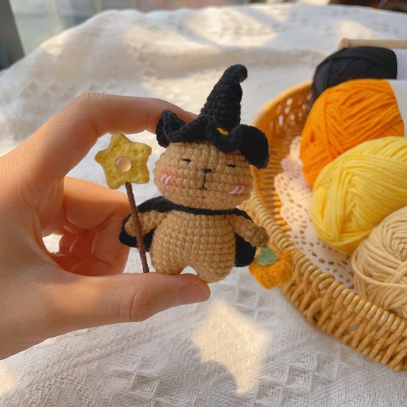 May include: A handmade crochet bunny wearing a black witch hat and cape, holding a star wand and a small pumpkin. The bunny is tan with pink cheeks. The background includes yarn in a basket.
