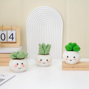 May include: Three crocheted succulent plants in white pots with smiling faces. The plants have green leaves and are arranged on a white surface. A wooden calendar and a white arched wall decor are in the background.