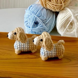 May include: Two handmade crochet dachshund dogs in tan and white with blue and white patterned sweaters. The dogs are on a wooden tray with balls of yarn in the background. The yarn colors are blue, tan, and white.