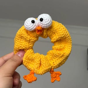 Könnte beinhalten: Ein handgefertigtes gelbes Häkel-Haargummi in Form eines Küken. Das Haargummi hat große weiße Augen mit schwarzen Pupillen, einen orangefarbenen Schnabel und orangefarbene Füße. Das Haargummi wird von einer Hand gehalten.