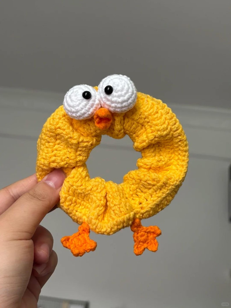 May include: A handmade yellow crochet hair scrunchie shaped like a chick. The scrunchie features large white eyes with black pupils, an orange beak, and orange feet. The scrunchie is held by a hand.