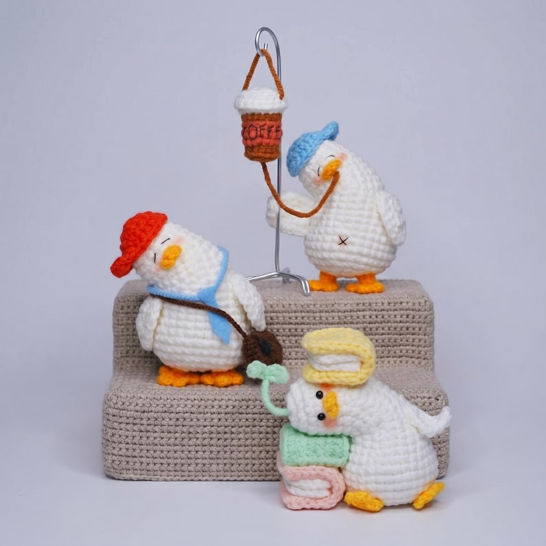 May include: Three white crocheted duck figures. One wears a red cap and blue tie with a brown bag. Another stands under a metal stand holding a brown crocheted coffee cup with the word COFFEE. The third rests on pastel books with a green sprout.