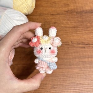 May include: A white crocheted bunny wearing a pink dress and a flower crown with pink, yellow, and blue flowers. The bunny has black eyes and a small pink nose.