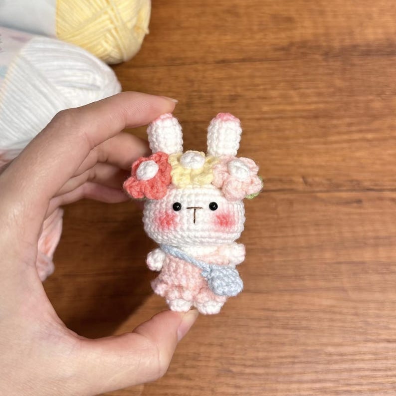 May include: A white crocheted bunny wearing a pink dress and a flower crown with pink, yellow, and blue flowers. The bunny has black eyes and a small pink nose.