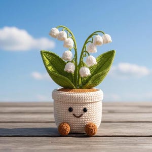 Op de afbeelding: Een gehaakte lelietje-van-dalen plant in een crèmekleurige pot met een lachend gezicht. De plant heeft witte klokvormige bloemen en groene bladeren. De pot heeft twee kleine bruine voetjes aan de basis en een textuur, wat zorgt voor een schattig uiterlijk.