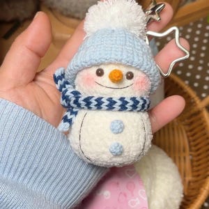 May include: A handmade snowman keyring with a blue knitted hat, scarf, and buttons. The snowman has a carrot nose, rosy cheeks, and a smiling mouth. A silver star-shaped charm is attached. The snowman is approximately 7.6 cm tall.