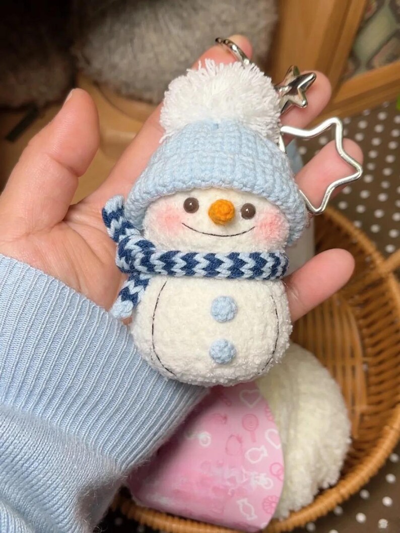 May include: A handmade snowman keyring with a blue knitted hat, scarf, and buttons. The snowman has a carrot nose, rosy cheeks, and a smiling mouth. A silver star-shaped charm is attached. The snowman is approximately 7.6 cm tall.