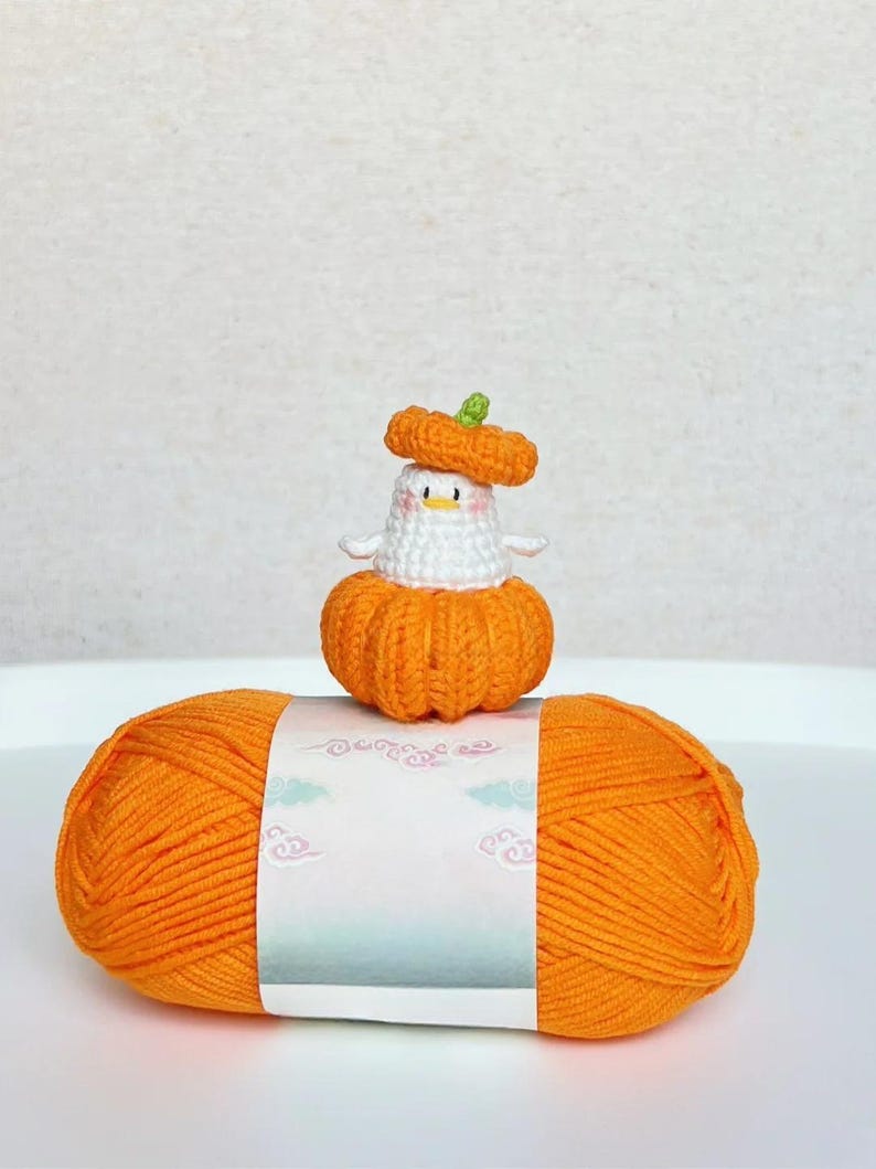 May include: An orange yarn ball with a white label featuring a decorative design. A small, handmade crochet duck wearing an orange pumpkin hat and body sits atop the yarn. The duck has a white body and a small green stem on its hat.