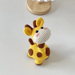 May include: A crocheted giraffe toy in yellow with brown spots and details. The giraffe has a white muzzle with pink cheeks and black eyes. The ears and small horns are brown. The toy is sitting on a white surface.