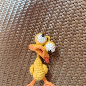 May include: A handmade, yellow crochet duck with orange feet, beak, and tongue. The duck has large, bulging eyes with red and white detailing. The whimsical design is set against a textured, brown background.
