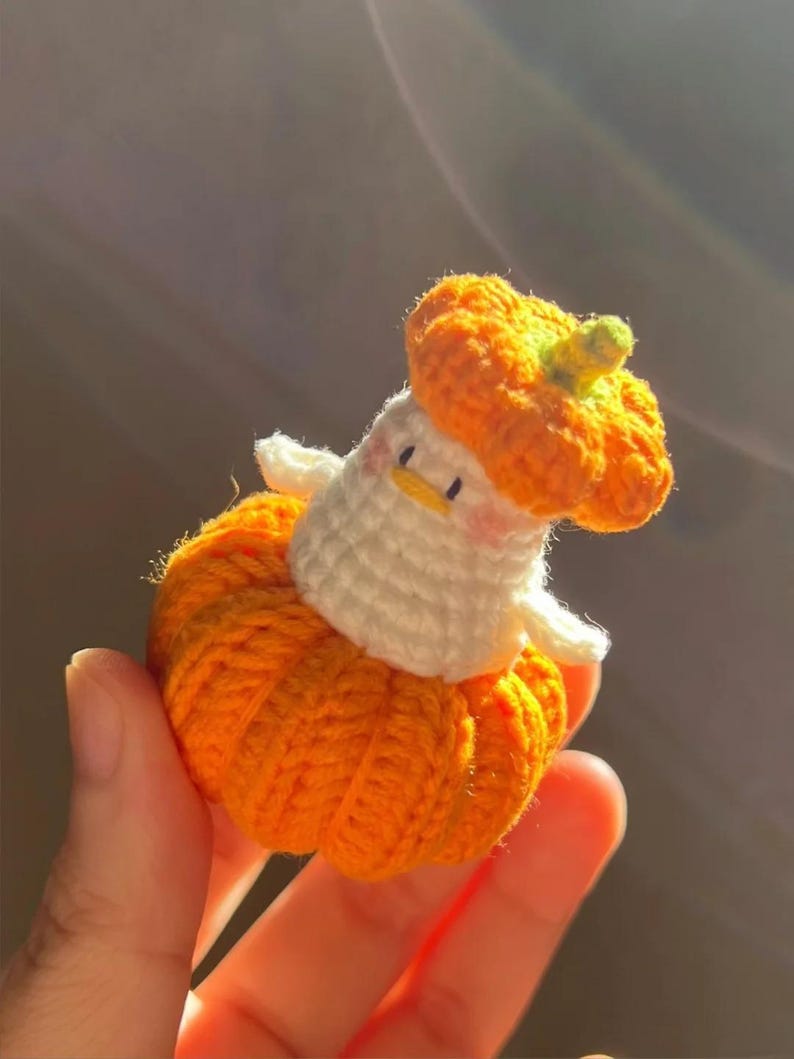 May include: A handmade crochet pumpkin-themed figure. The figure is primarily orange, with a white body and a small, yellow beak. The pumpkin hat has a green stem. The figure is held in a hand.