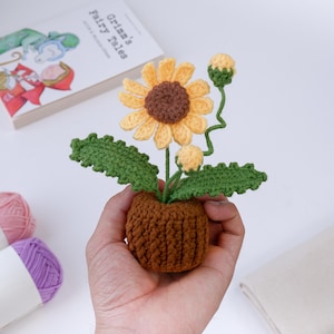 Könnte beinhalten: Eine handgefertigte gehäkelte Sonnenblume in einem braunen Topf. Die Sonnenblume hat gelbe Blütenblätter, eine braune Mitte und grüne Blätter. Der Topf ist ebenfalls braun. Die Blume wird in einer Hand gehalten, mit einem Buch mit dem Titel "Grimms Märchen" im Hintergrund.