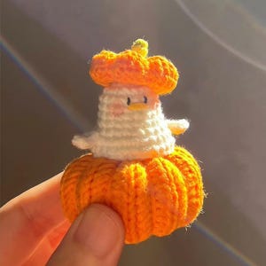 May include: A handmade crochet duck wearing a pumpkin hat and dress. The duck is white with a yellow beak and small black eyes. The pumpkin hat and dress are bright orange, with a green stem on the hat. The duck is held in a person's hand.