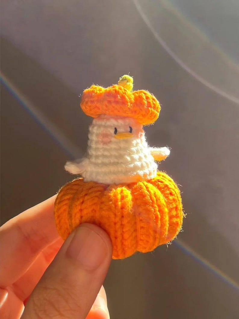 May include: A handmade crochet duck wearing a pumpkin hat and dress. The duck is white with a yellow beak and small black eyes. The pumpkin hat and dress are bright orange, with a green stem on the hat. The duck is held in a person's hand.