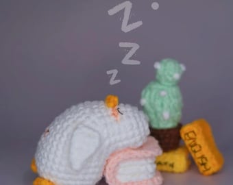 May include: A crocheted white sleeping duck with yellow feet and beak, resting on a pink and white pillow. A green cactus and two yellow blocks with text are nearby. The word "Z" is written above the duck.