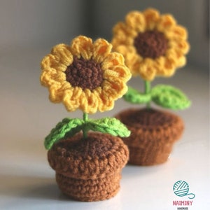 Puede incluir: Dos girasoles de crochet en macetas marrones con hojas verdes. Los girasoles son amarillos con centros marrones.