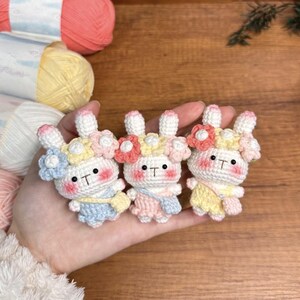 May include: Three crocheted bunny plushies with flower crowns. The bunnies are wearing different colored outfits: blue, pink, and yellow.