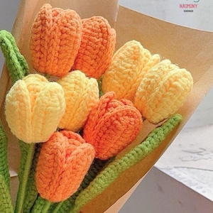 May include: A bouquet of crocheted tulips in shades of orange and yellow, with green crocheted stems. The bouquet is wrapped in brown paper and tied with a green ribbon. The label on the paper reads "NAIMINY HANDMADE".