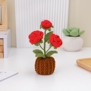 May include: A handcrafted crochet flower arrangement featuring three vibrant red roses with green leaves and stems, nestled in a brown pot. The handmade floral decor adds a touch of charm to any space.