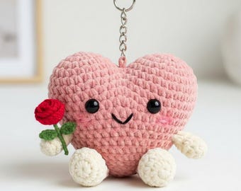 May include: A crochet heart-shaped keyring in pink yarn. The heart has black button eyes, a stitched smile, and rosy cheeks. It holds a red rose and has white, round feet. A silver keyring is attached.