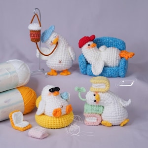May include: Four white crocheted duck figures with orange feet. One wears a blue cap and holds a coffee cup, one rests on a blue sofa wearing a red cap, one wears black sunglasses in a yellow float, and one sits with stacked books and a green sprout.