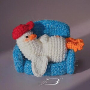 May include: A white crocheted duckling with a red beret and orange feet, resting on a small blue crocheted armchair. The duckling has black bead eyes and a small orange beak. A white line drawing of a smartphone is positioned below the armchair.