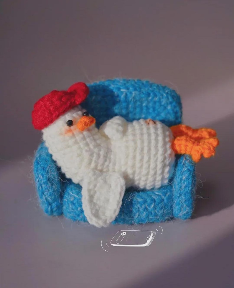 May include: A white crocheted duckling with a red beret and orange feet, resting on a small blue crocheted armchair. The duckling has black bead eyes and a small orange beak. A white line drawing of a smartphone is positioned below the armchair.