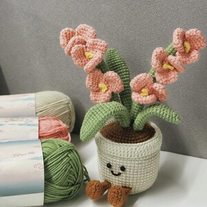 Puede incluir: Una maceta de flores de ganchillo con una cara sonriente, que sostiene un ramo de flores rosas con centros amarillos y hojas verdes. La maceta es de color crema, con tierra marr&oacute;n y dos pies marrones. Se ven madejas de hilo en verde, rosa y crema.