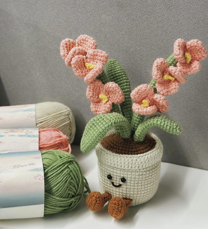 Puede incluir: Una maceta de flores de ganchillo con una cara sonriente, que sostiene un ramo de flores rosas con centros amarillos y hojas verdes. La maceta es de color crema, con tierra marr&oacute;n y dos pies marrones. Se ven madejas de hilo en verde, rosa y crema.