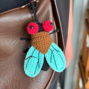 May include: A crocheted insect keyring featuring two large red eyes, a brown textured head and body, and two light blue wings with black stitched vein details. The insect is attached to a black cord and a metal ring for hanging on a bag.