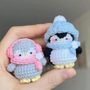 以下が含まれることがあります： 手作りの編みぐるみペンギンの置物2つ。1つは水色の帽子とスカーフ、もう1つはピンクのイヤーマフとスカーフを着用。どちらも白黒の体、黄色の足、ピンクの頬をしています。冬の装飾に最適です。