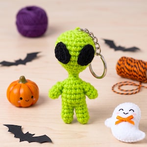 May include: A lime green crocheted alien keychain featuring a large head with two oval black stitched eyes, a slender neck, and a small body attached to a silver metal split ring. The alien is crafted with visible yarn stitches.