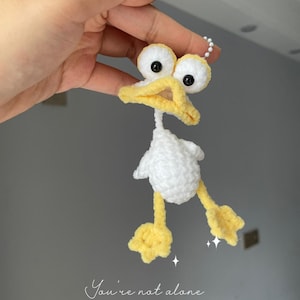 May include: A handmade crochet duck keychain with a white body, yellow beak and feet, and black eyes. The duck is held up by a hand. The text "You're not alone" is written in cursive below the duck.