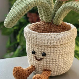 Puede incluir: Una planta de ganchillo en una maceta blanca con una cara sonriente y piernas marrones. La planta tiene tallos verdes y flores rosas. La maceta est&aacute; llena de hilo marr&oacute;n, que se asemeja a tierra. El fondo est&aacute; borroso, con follaje verde.